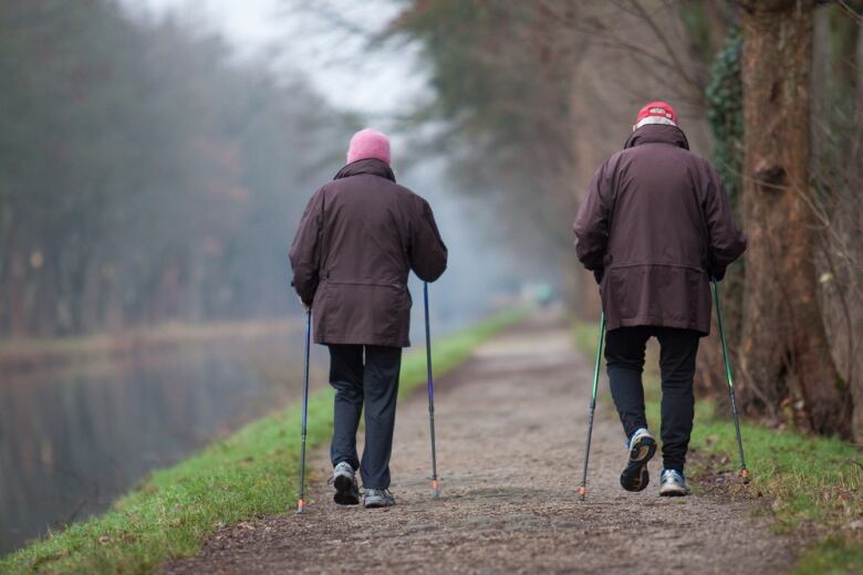 Two seniors use walking sticks to walk down a forest path.