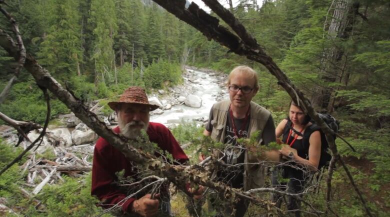 Three people examine a tree branch in a wilderness area while the white water of a forest stream flows swiftly behind them.