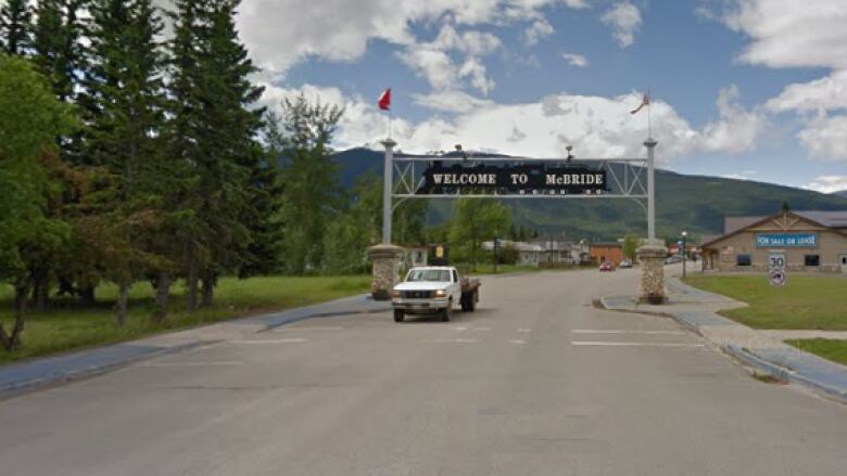 A white pick up truck drives along a highway underneath a sign that arches over the road ans says welcome to McBride.