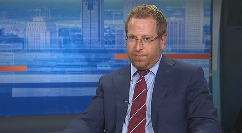 A man in a suit sits in front of a backdrop showing a city skyline in a TV studio.