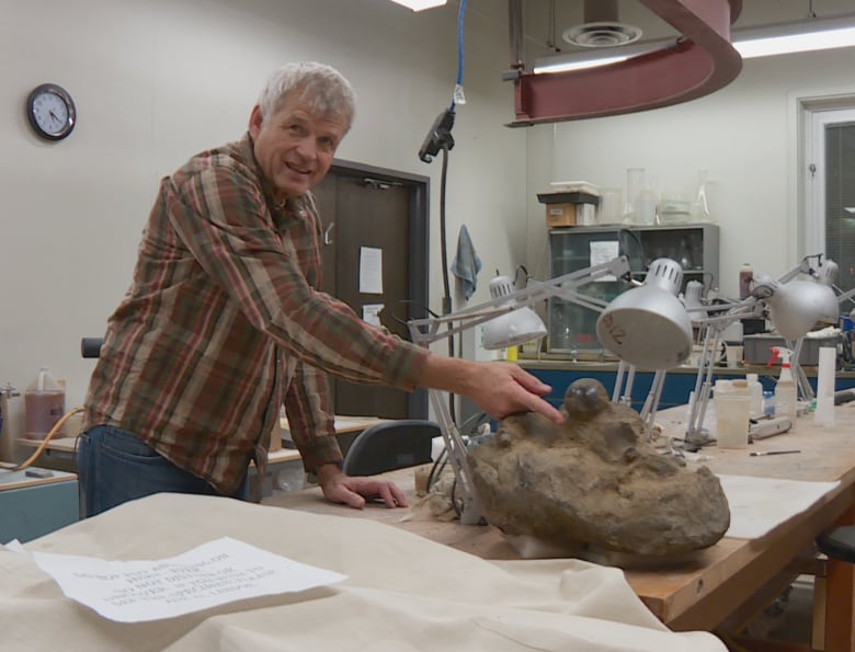 A white man wearing jeans and a flannel dress shirt is standing in a research lab, pointing to a fossil on his desk.