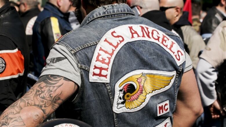 The back of a hells angels jacket is pictured in a close up photo worn by a man with big tattooed arms 