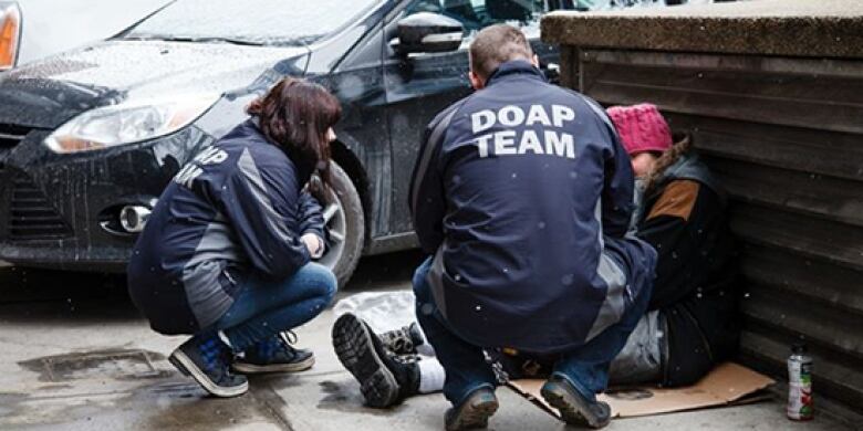 Two people with the words DOAP TEAM written across their jackets are crouched down next to someone sitting on the ground against a an air vent. None of the people's faces is visible.