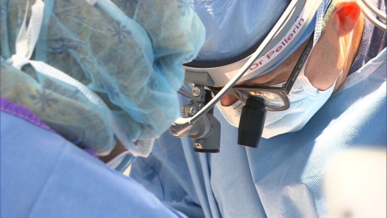 A surgeon in blue scrubs with a magnifying loupe headset works on a patient.