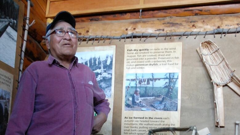 An elder stands in front of a moose hide display in a wooden cabin.