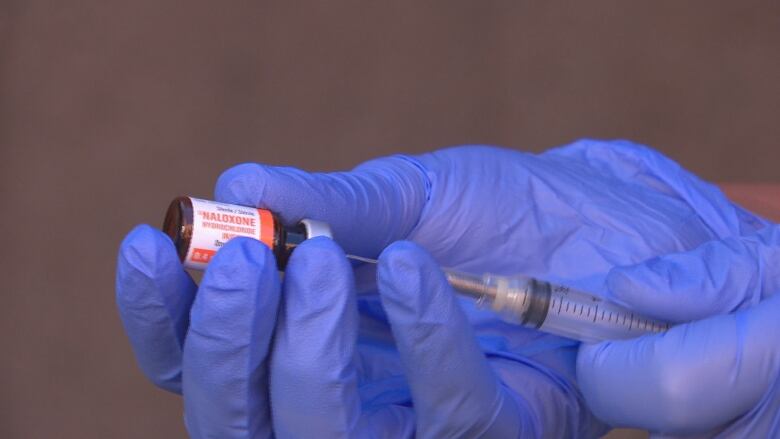 A closeup shot of two hands in purple medical gloves holding a syringe that says naloxone.