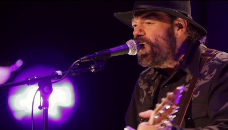 A man sings into a microphone and plays guitar in a dark room.