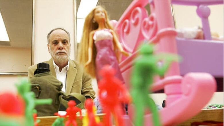 A man with a white beard sits in a tan blazer behind what appears to be toys.