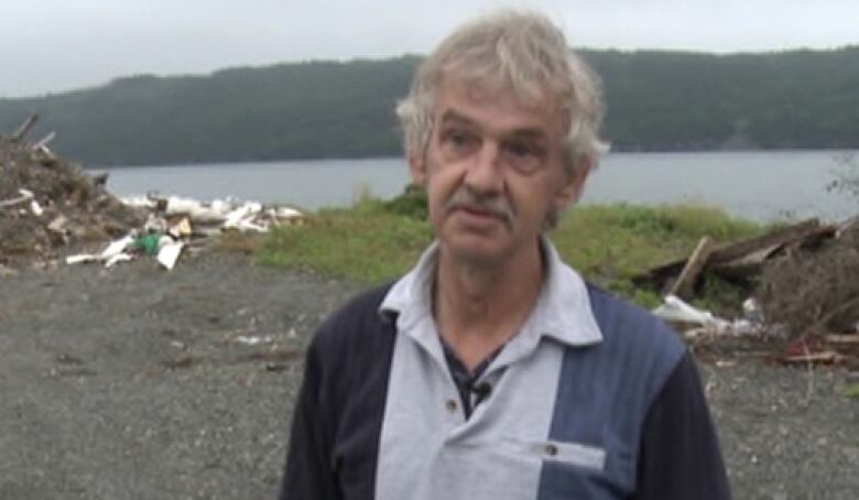 A thin grey-haired man is in the foreground. He is standing in front of a body of water. 