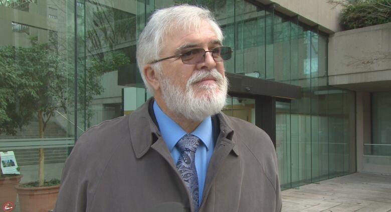 A white man wearing glasses and a black coat speaks in front of a courthouse.