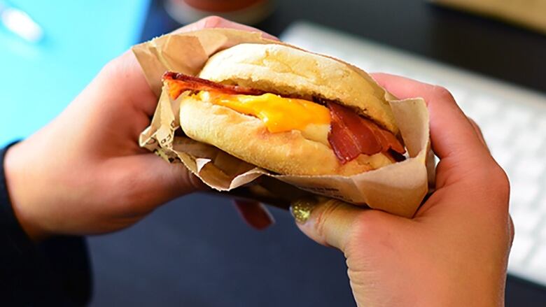 A pair of hands holds a breakfast sandwich with an A&W cup and bag and a computer keyboard in the background.