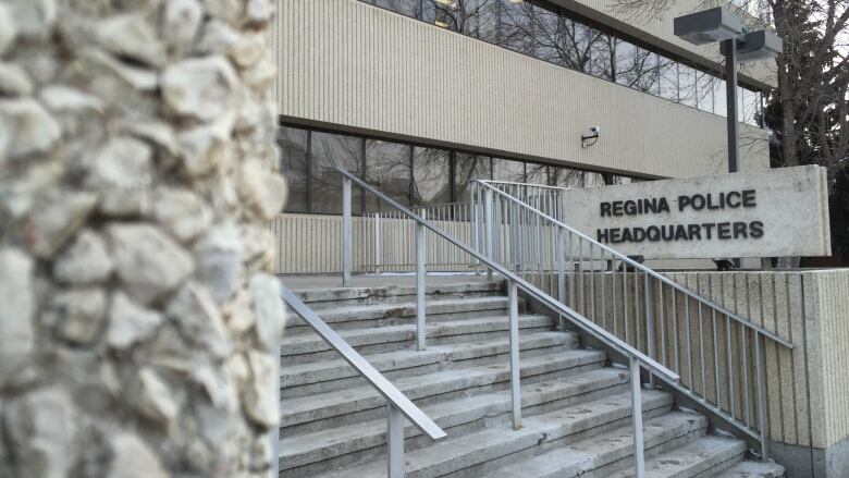 A sign in front of a stone building reads, 'Regina Police Headquarters.' There are stairs with metal rails in front of the sign.