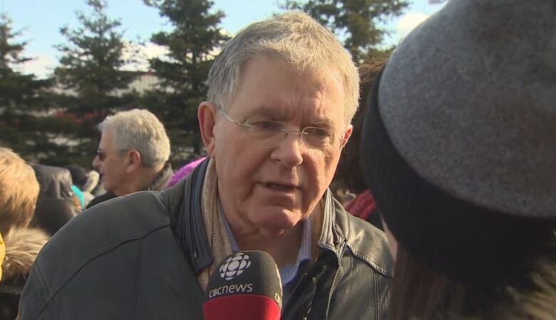 An older man wearing a leather jacket speaks in front of a microphone.