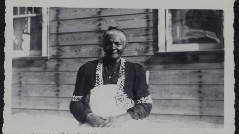 An archival black and white portrait of Mattie Mayes sitting in front of a wooden building.