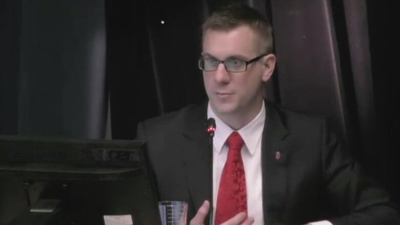 A man wearing glasses is wearing a black suit and red tie. He is sitting in front of black curtains. A thin black microphone is in front of him. 