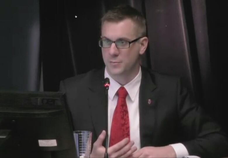 A man wearing glasses is wearing a black suit and red tie. He is sitting in front of black curtains. A thin black microphone is in front of him. 