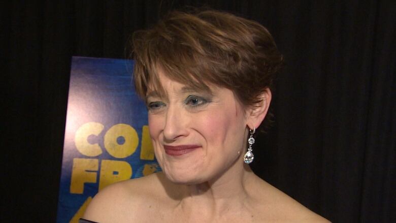 A woman wearing a gown and earrings stands in front of a show poster that says Come From Away. 