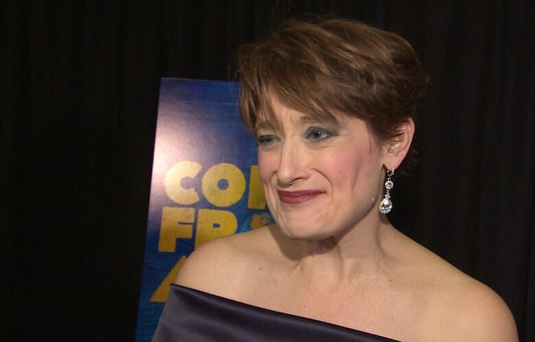 A woman wearing a gown and earrings stands in front of a show poster that says Come From Away. 