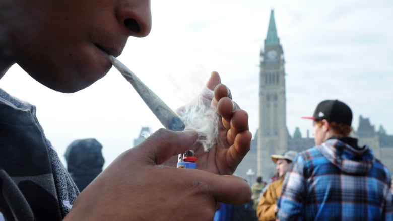 A person lights a marijuana joint as others gather in the background. The Peace Tower of Parliament Hill can be seen in the background.