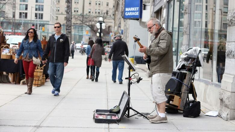 A man wearing a green jacket and white shorts plays an electric guitar on an Ottawa street. A couple walk by him on the left of the photo