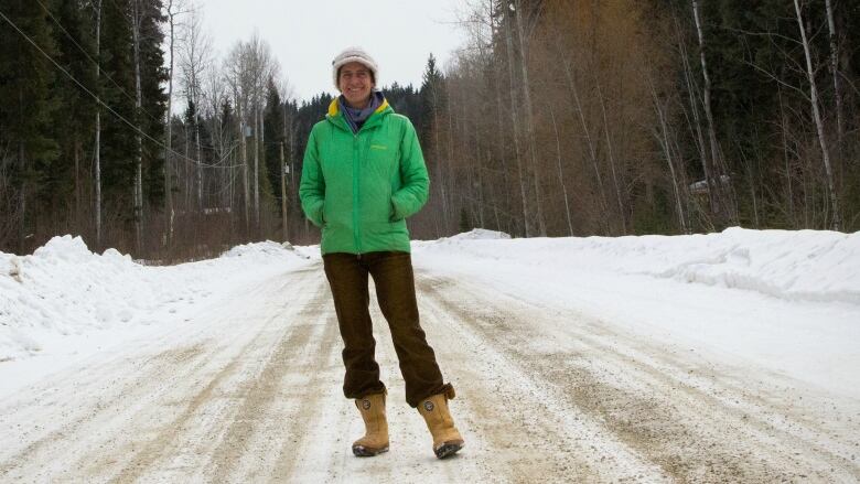 A woman stands in the middle of a snowy rural road, wearing a white hat, green coat, and brown pants and snow boots. 