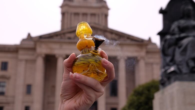 A hand holding a bong in front of the Manitoba Legislative Building.