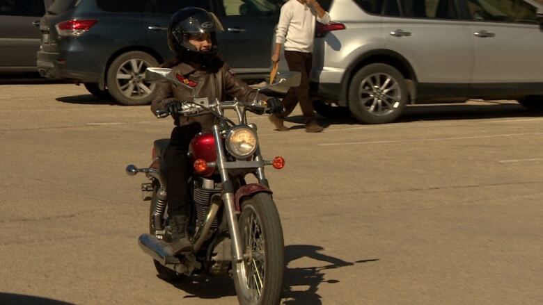 A woman wearing a helmet and leather jacket rides a motorcycle through a parking lot.