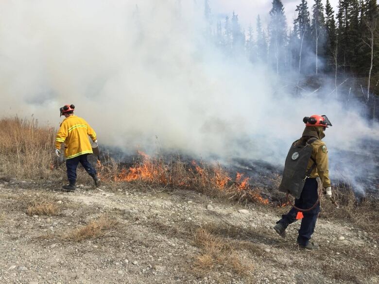 Firefighters walking beside a small grass fire, with smoke billowing into the sky.