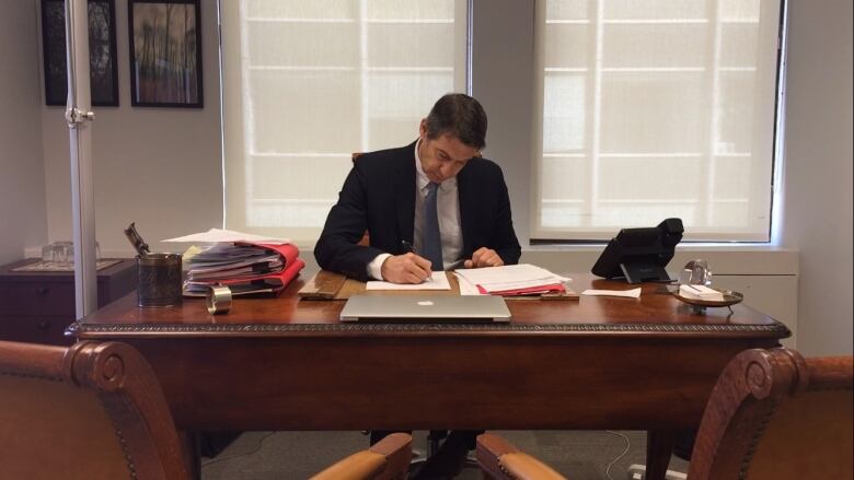 A man sits behind a wooden desk and writes on a paper. 