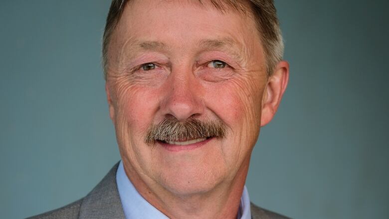 A portrait of a middle-aged man with a mustache, wearing a grey suit with a medal on the lapel, a light blue shirt and dark blue patterned tie.