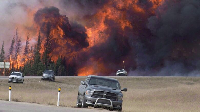 An enormous wall of fire and smoke is seen in the background behind vehicles on a highway.