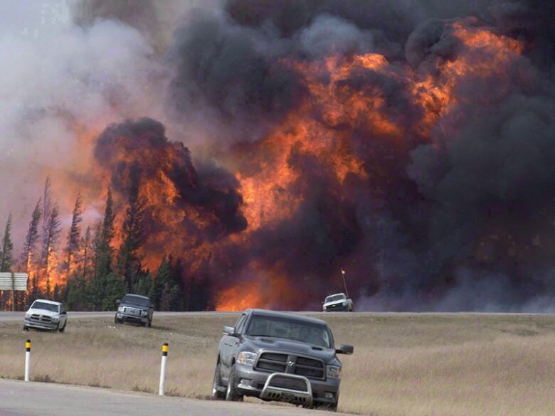 An enormous wall of fire and smoke is seen in the background behind vehicles on a highway.