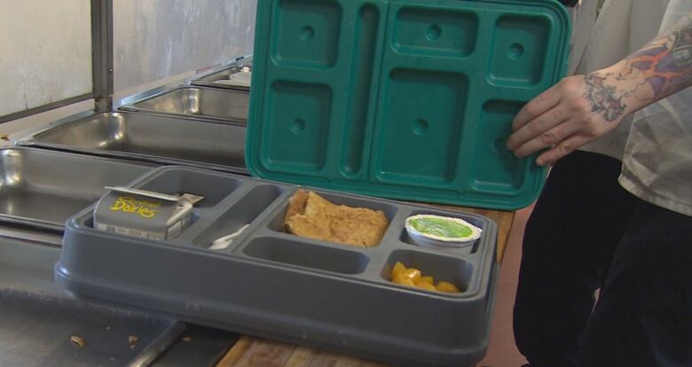 Milk, juice and bread sit on a grey tray. A tattooed hand is holding onto a green lid hovering over the tray. 