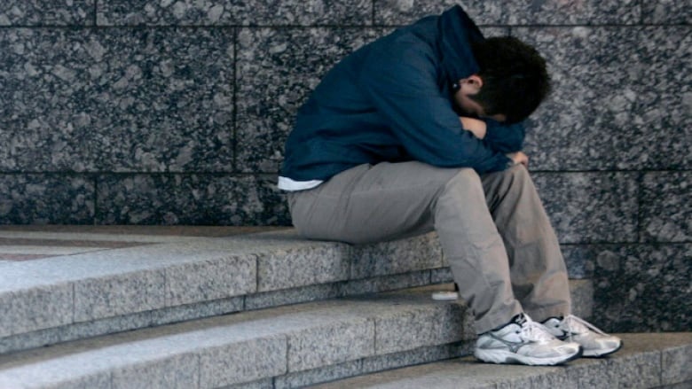 A male teen sits atop a curved stone staircase next to a stone wall, with his head curled down into his arms and his face hidden.