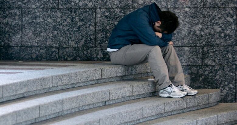 A male teen sits atop a curved stone staircase next to a stone wall, with his head curled down into his arms and his face hidden.