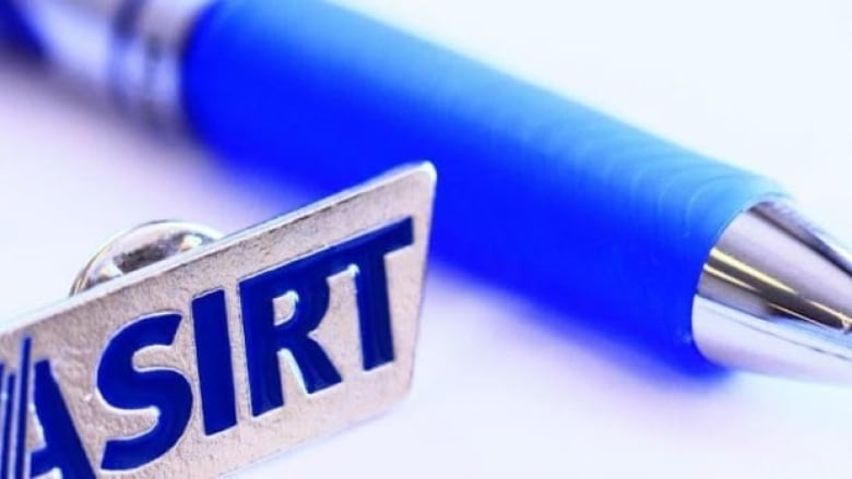 A close up image of a silver pin that says ASIRT in bold, blue lettering. A pen is seen beside the pin. 