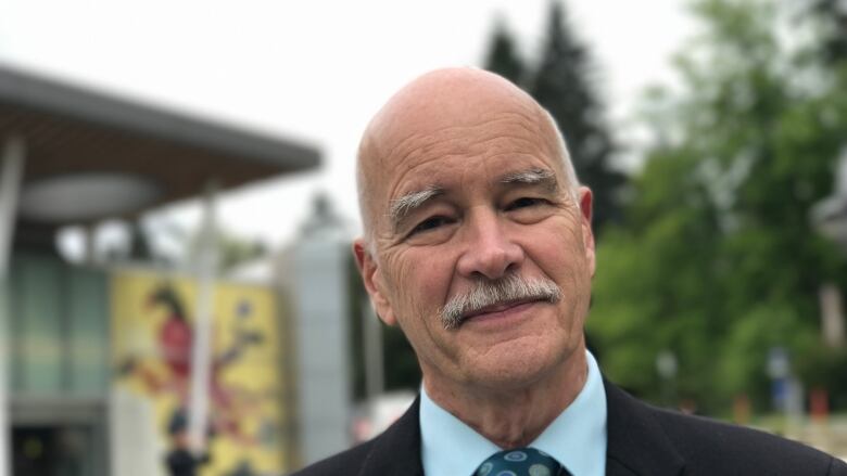 A bald man with a grey mustache is smiling. He's wearing a suit with a blue shirt and tie. 