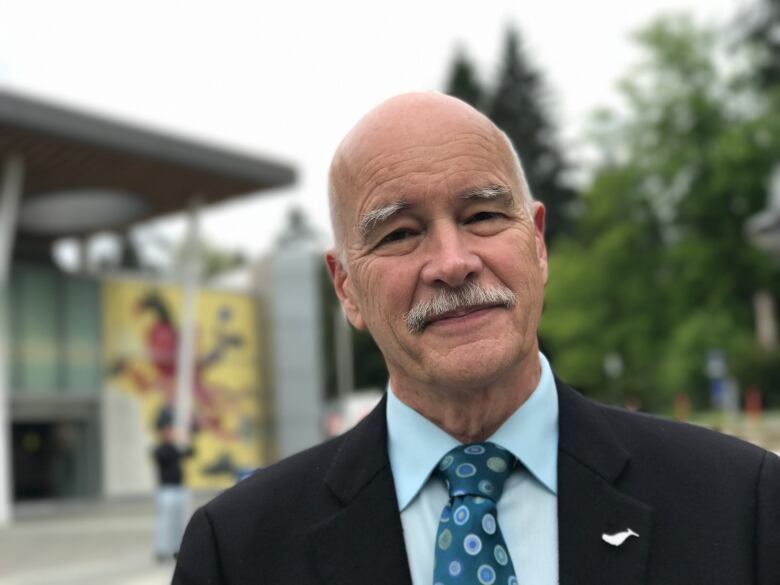 A bald man with a grey mustache is smiling. He's wearing a suit with a blue shirt and tie. 