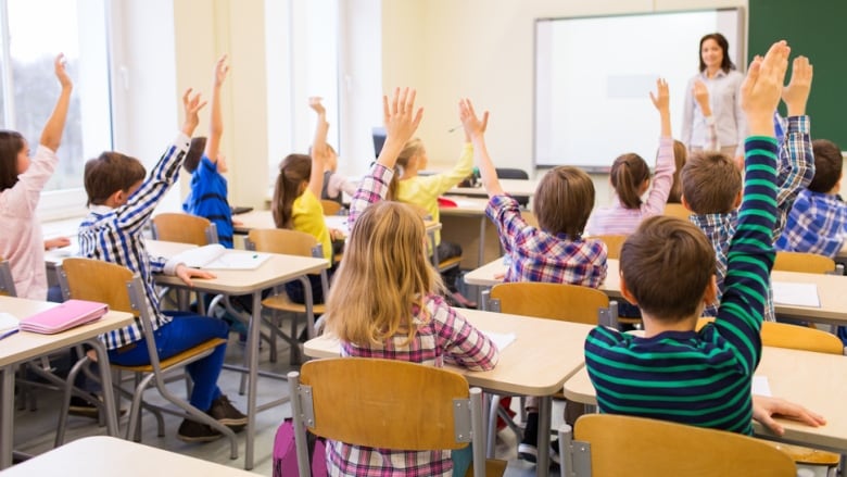 A female teacher stands at the front of a classroom as several students seated at their desks raise their hand.