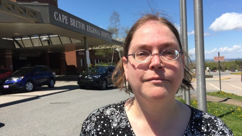 A woman with her hair pulled back, wearing glasses and a black-and-white print blouse, stands in front of the entrance to a hospital.