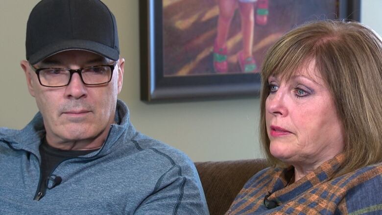 A man and a woman with serious expressions sit on a couch in a home.