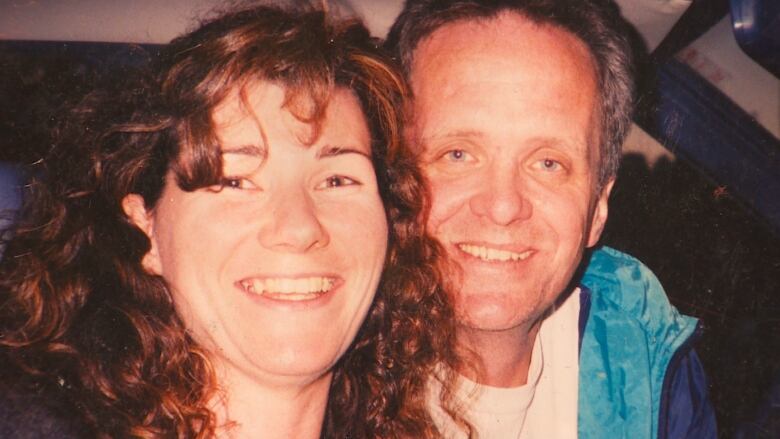 A woman and man smile at the camera from the inside of a car. Sasha Wood, left, has long, dark curls, and is seated closely next to her husband, Murray Shaw, who has grey hair, wearing a white t-shirt and a light blue windbreaker.