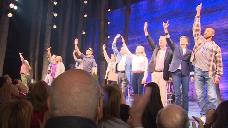 A cast of actors on a stage wave at audience.