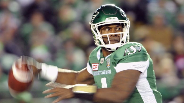 A man in a green and white jersey and football helmet pulls his arm back to fire a football. 