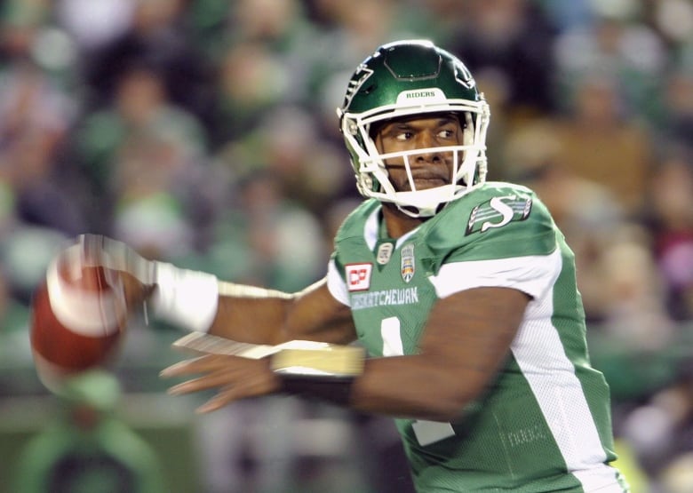 A man in a green and white jersey and football helmet pulls his arm back to fire a football. 