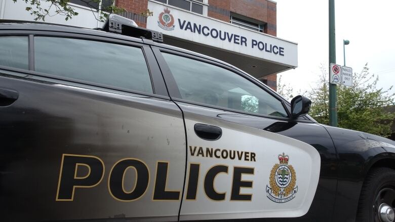 A closeup of a Vancouver police cruiser with its side door saying Vancouver Police in black lettering on a white background with the Vancouver police logo on it. The Vancouver police station is in the background.