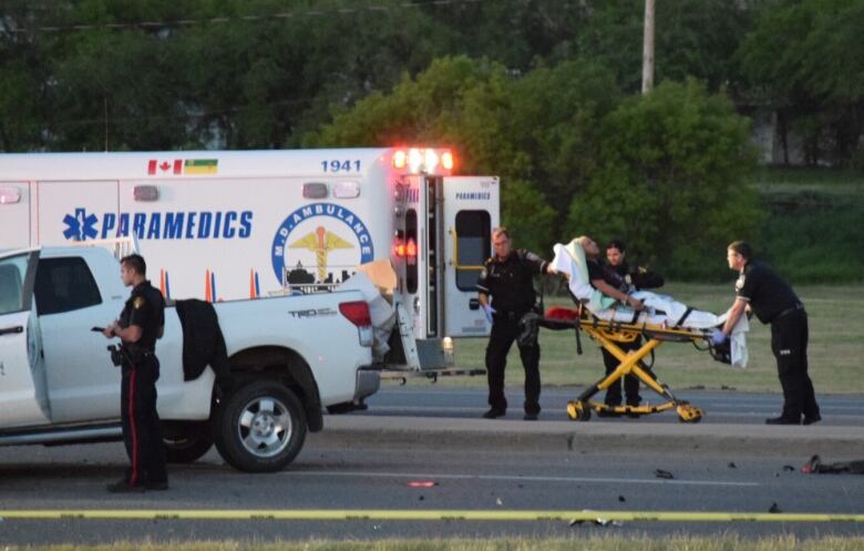 Two paramedics load a person on a gurney into the back of an ambulance at the scene of vehicle crash on a street.