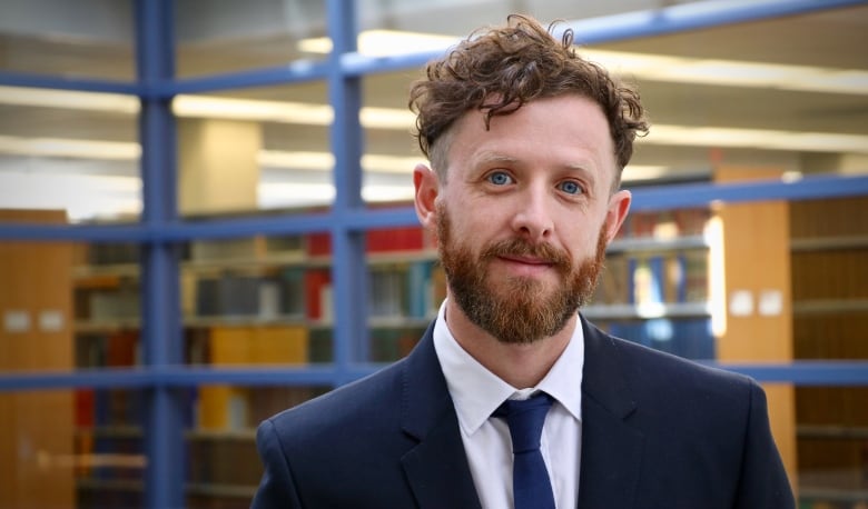 A man with brownish-red hair and a beard, wearing a navy jacket and tie, stands near bookshelves.