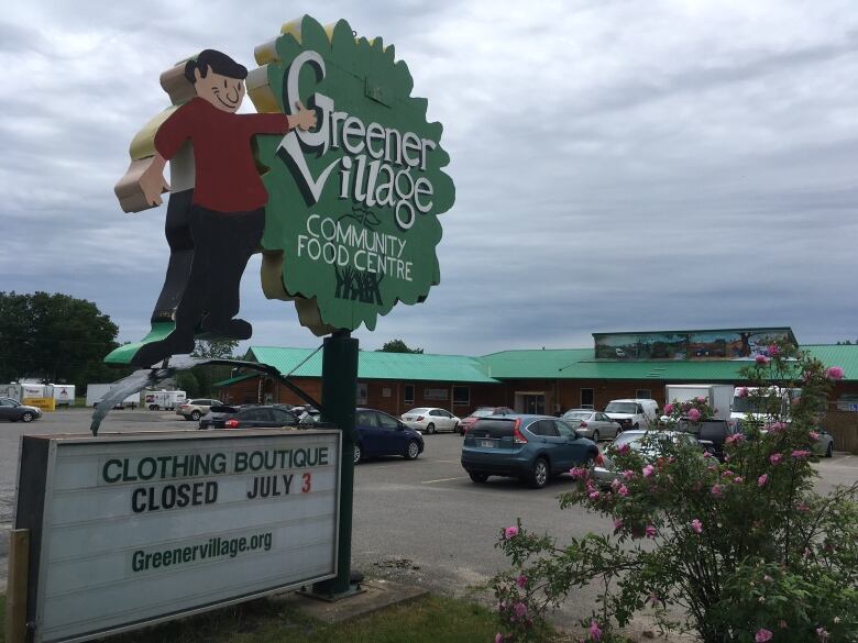 A green, circular sign next to a waving man statue saying Greener Village in front of the storefront in Fredericton