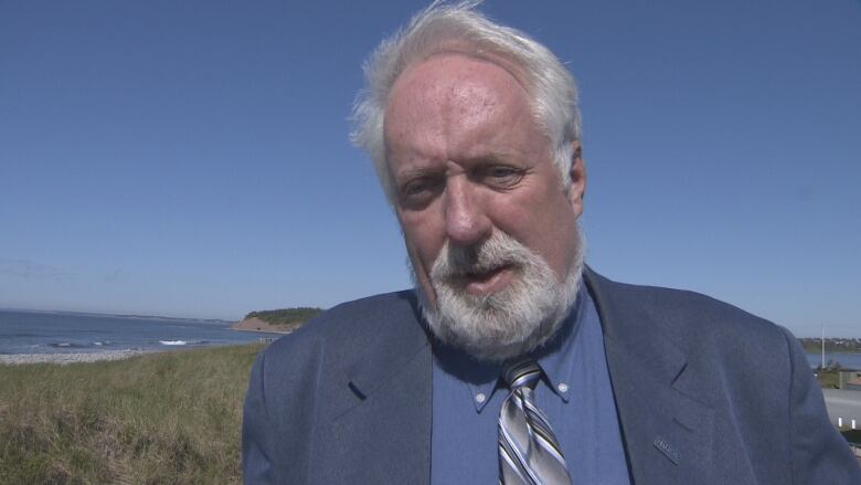 A white man with a blue shirt, navy jacket and tie stands outside in a coastal area. Blue water is visible behind him